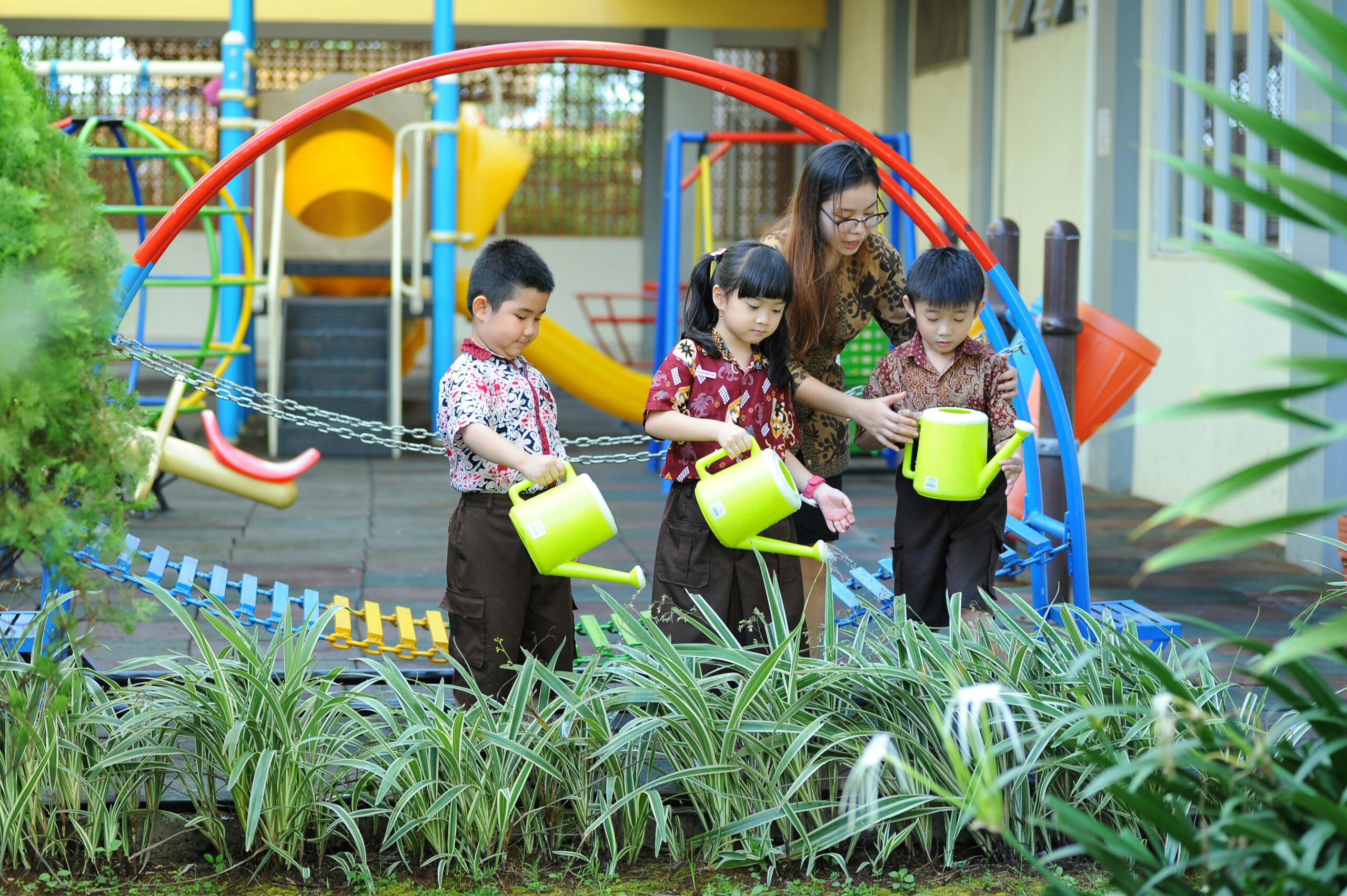 Children with a teacher learning to water plants in a colorful playground setting.