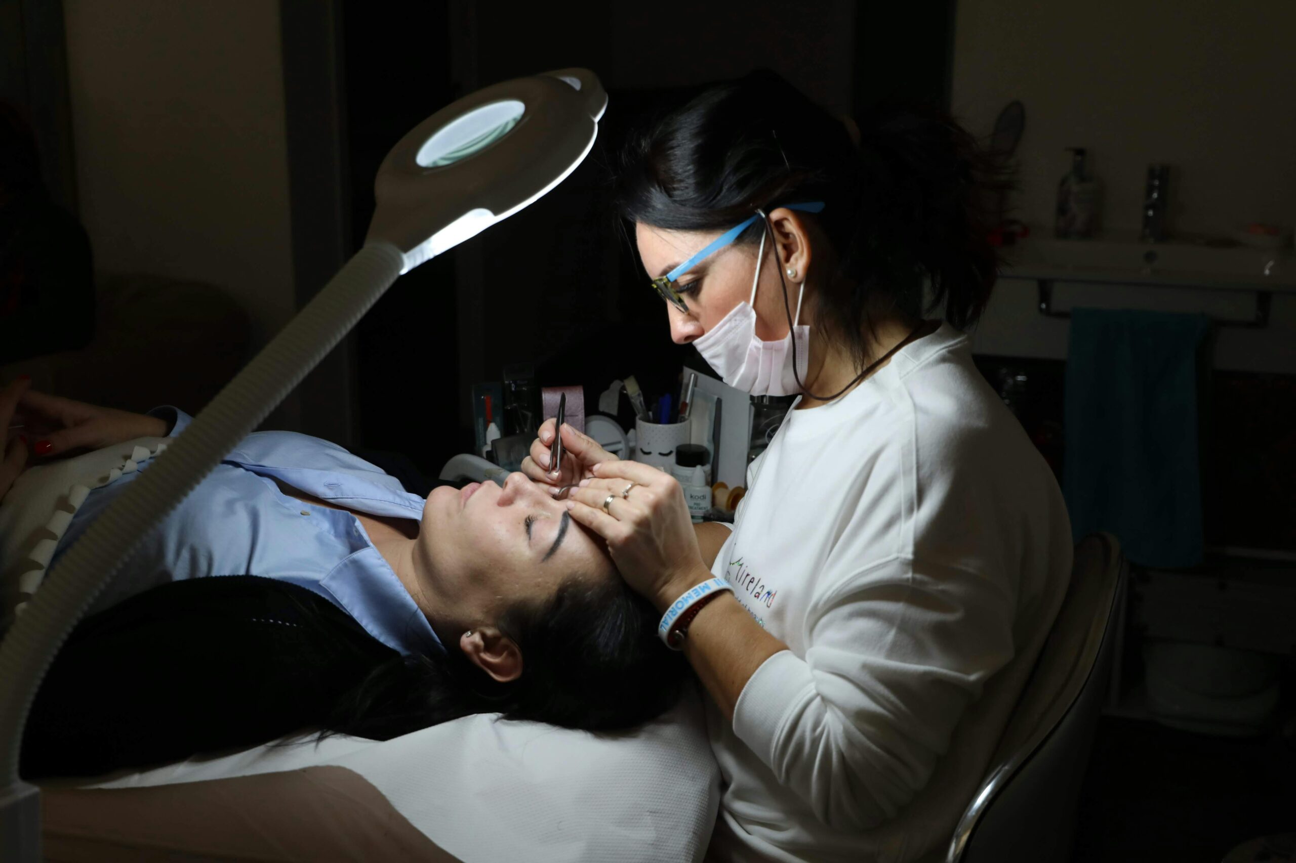 A beautician provides professional eyebrow care to a client in a salon, under focused lighting.