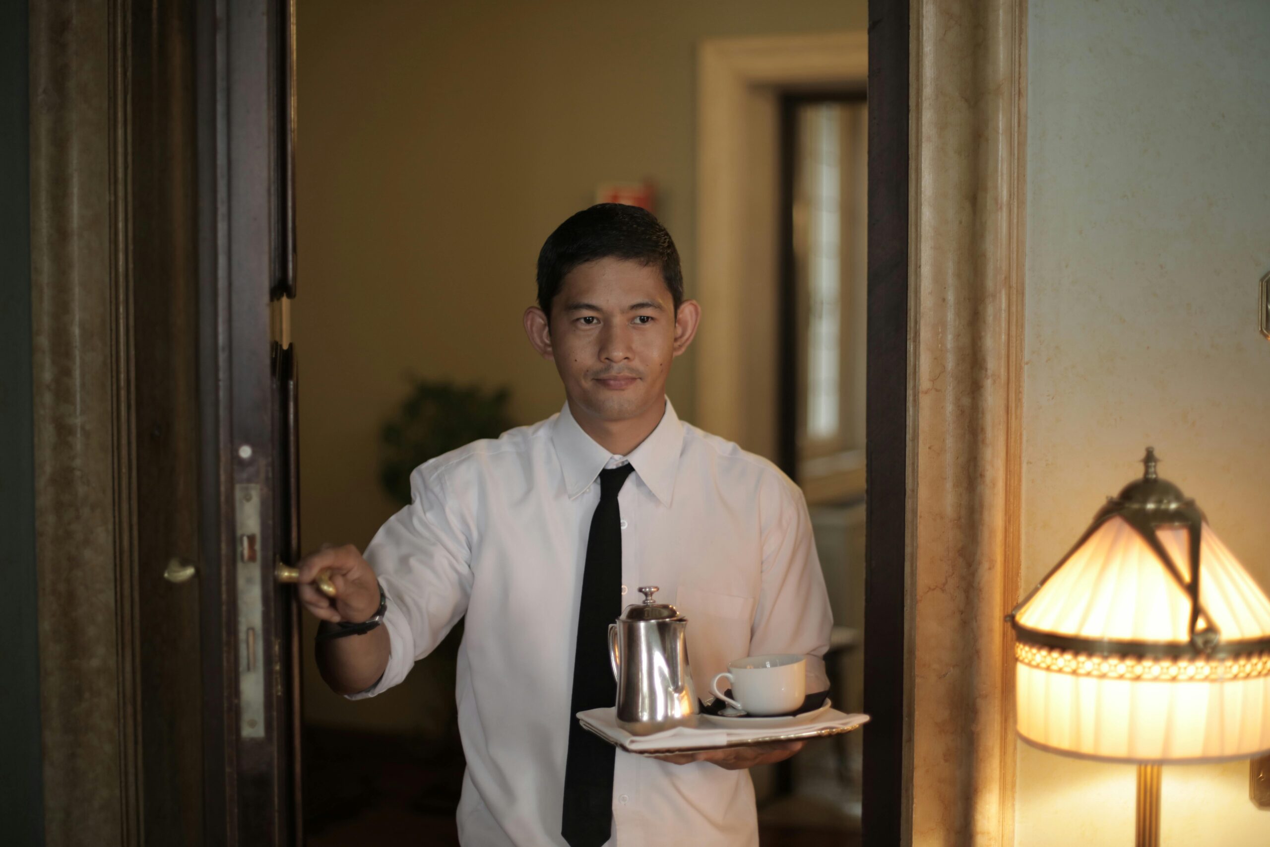 Positive ethnic male employee carrying tray with coffee while serving guest in cozy hotel room