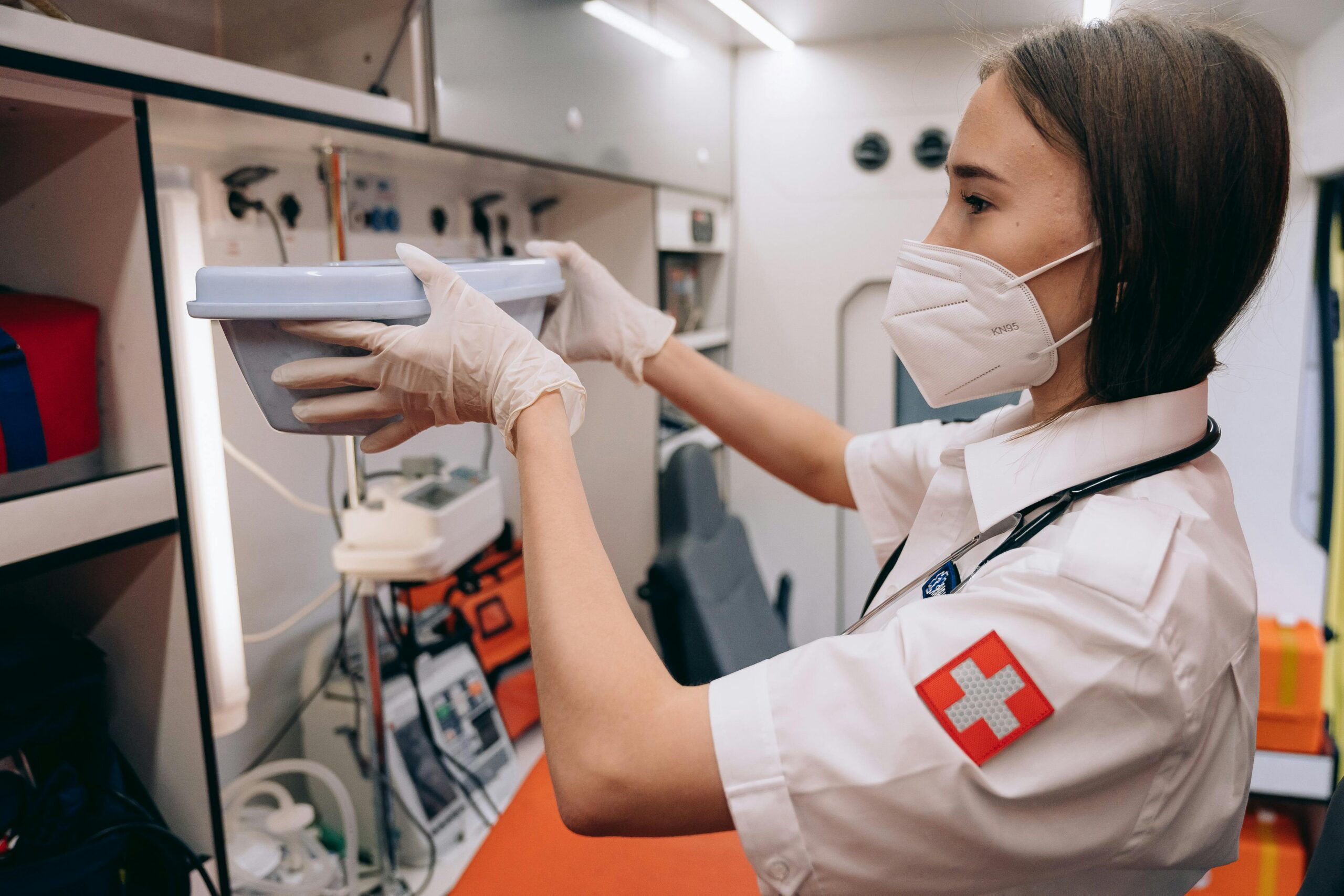 A female paramedic organizes medical supplies inside an ambulance, wearing a face mask and gloves.