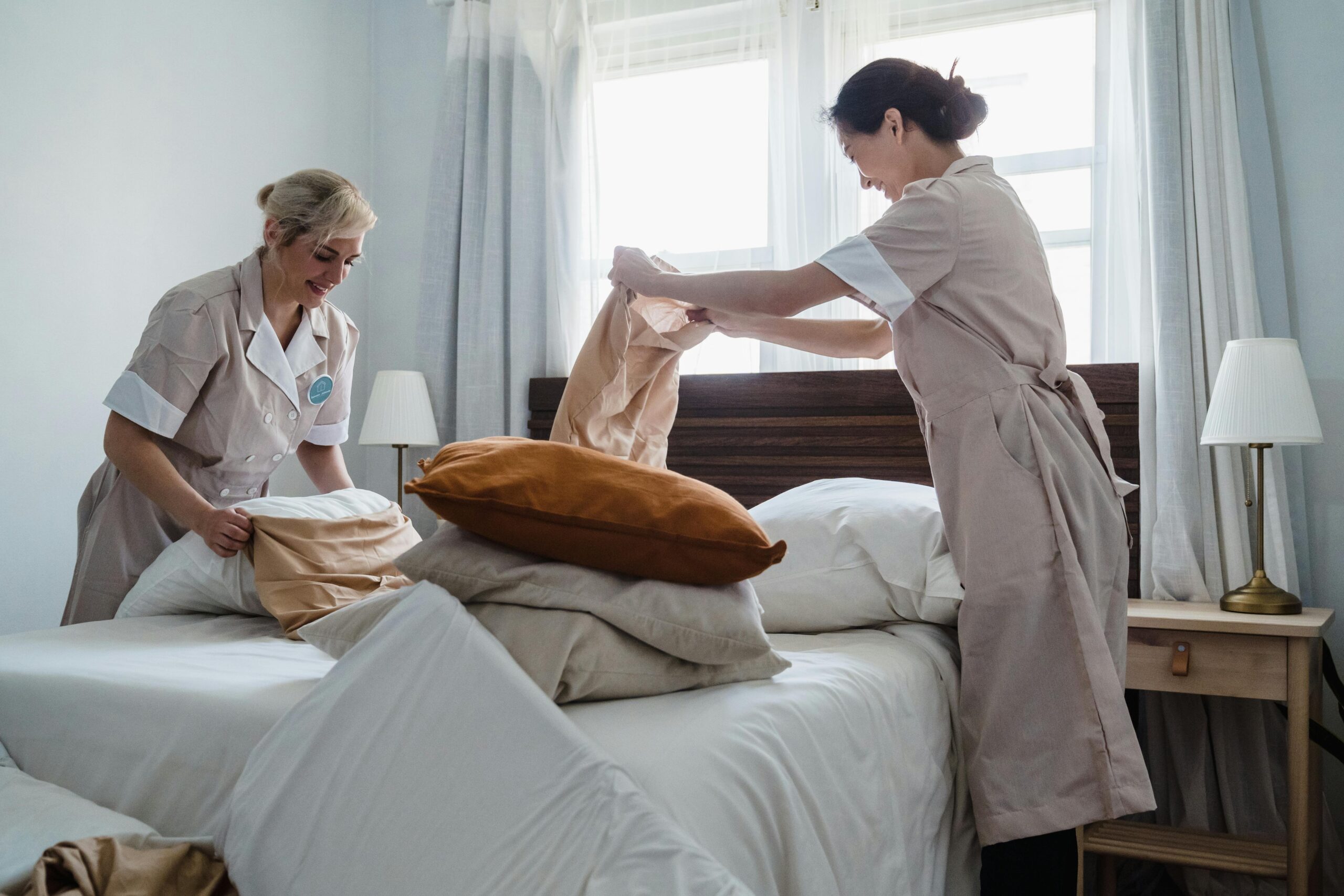 Two housekeepers in uniform fixing a hotel room bed with fresh linen and pillows.